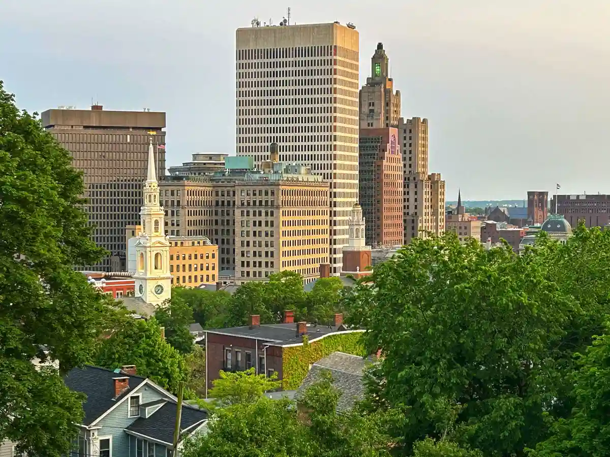 Downtown Providence, RI skyline with church steeple, high-rise buildings, and tree-lined residential rooftops in foreground