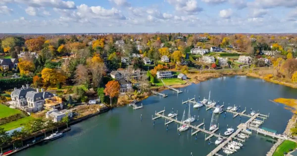 Aerial view of a coastal marina with docked sailboats, waterfront homes, and autumn foliage along the shoreline