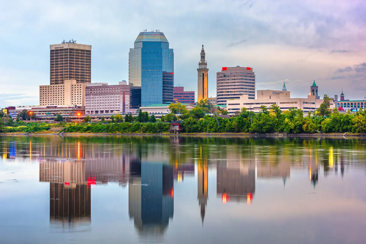 City skyline with tall buildings reflected in a calm river at dusk.