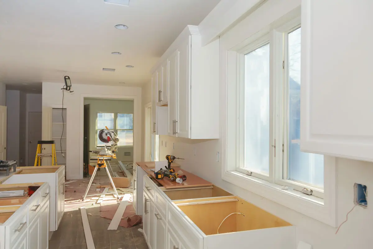 In-progress kitchen remodel in Tolland County, CT with white cabinetry installed, an unfinished cabinet drawer without face or hardware, a miter saw staged in the adjoining room, and tools and floor protection visible during construction